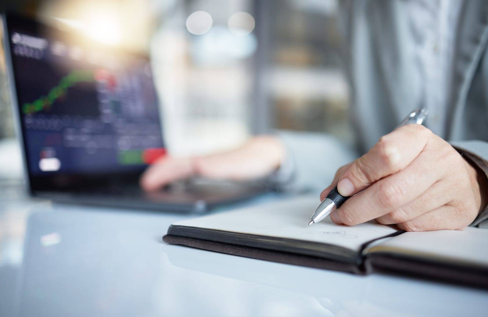 Trader, Hands And Writing Notes In Book By Laptop On Stock Market Chart Or Graph Statistics For Trading At The Office.