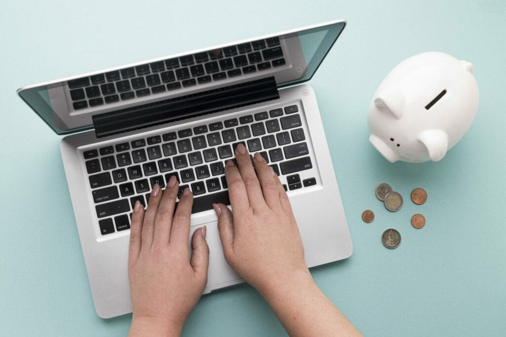 A hand operating a laptop for stock trading and a piggy bank on the side with coins scattered.